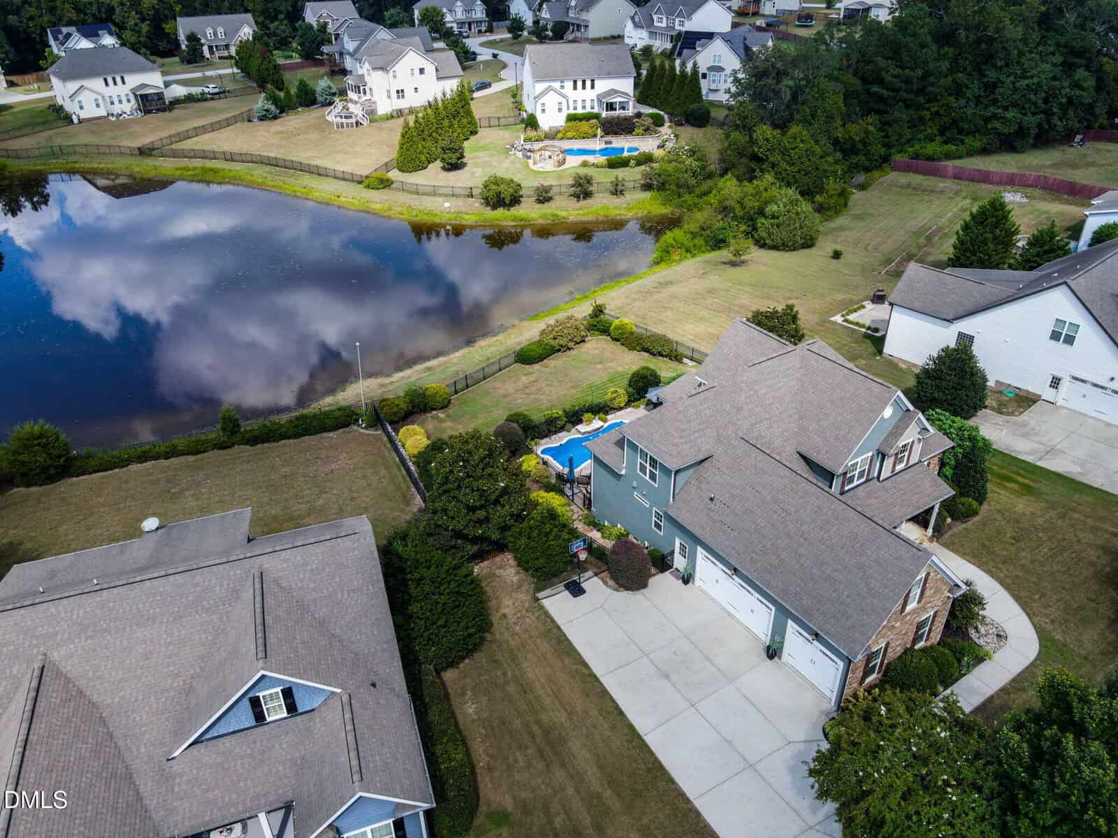 Aerial view of suburban homes with large yards, a backyard pool, and a small lake reflecting clouds and sky, surrounded by trees and neatly trimmed lawns.