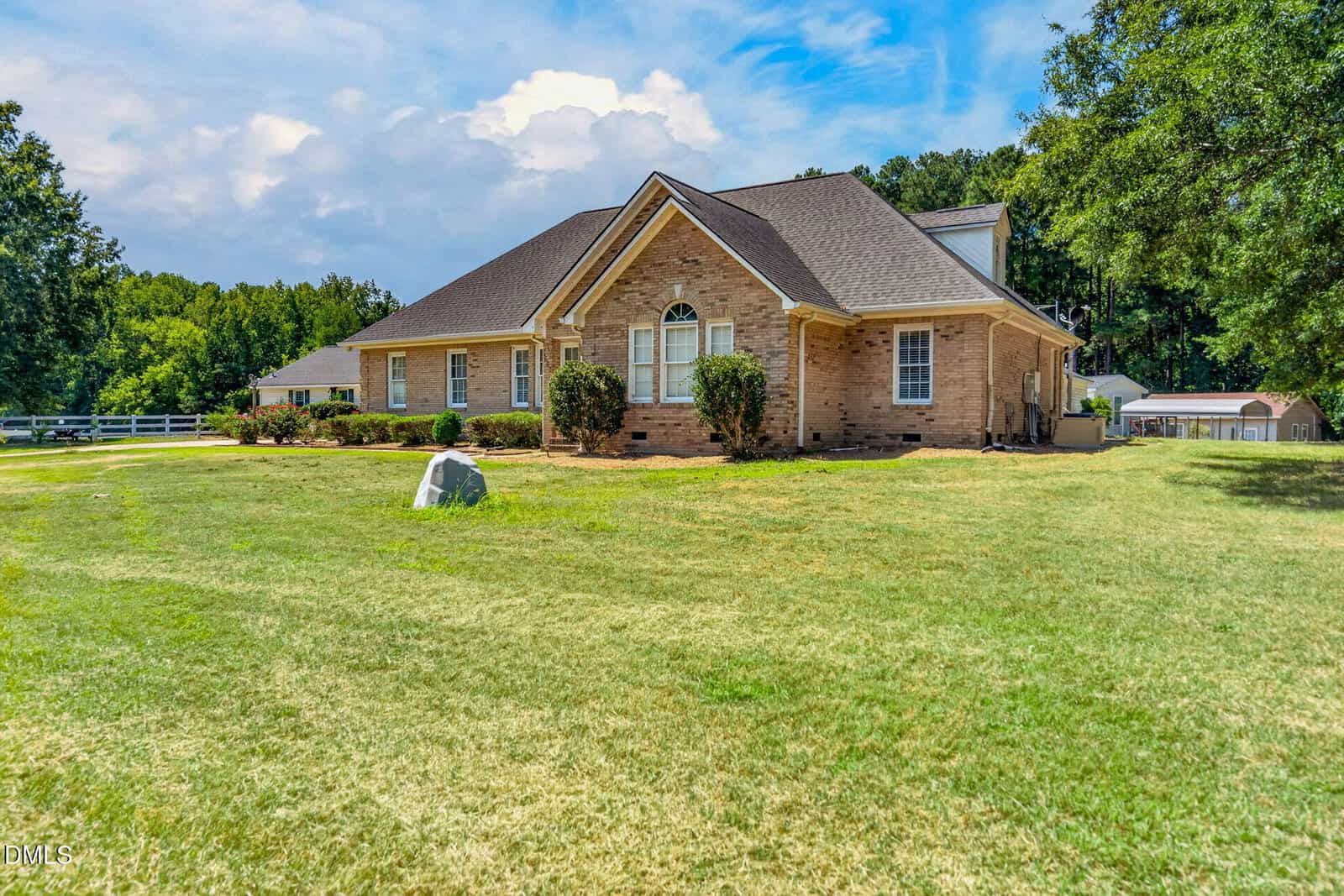 A one-story brick house with a gabled roof sits on a large grassy yard, surrounded by trees and a white fence, under a partly cloudy sky.