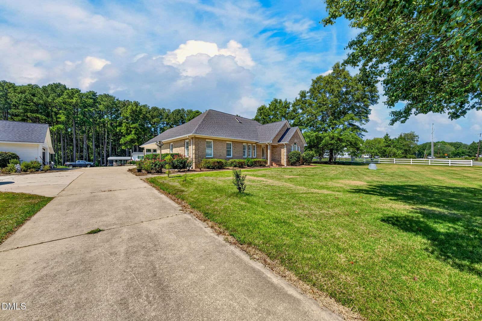 Single-story brick house with a gray roof, large front yard, concrete driveway, and trees in the background under a partly cloudy sky.
