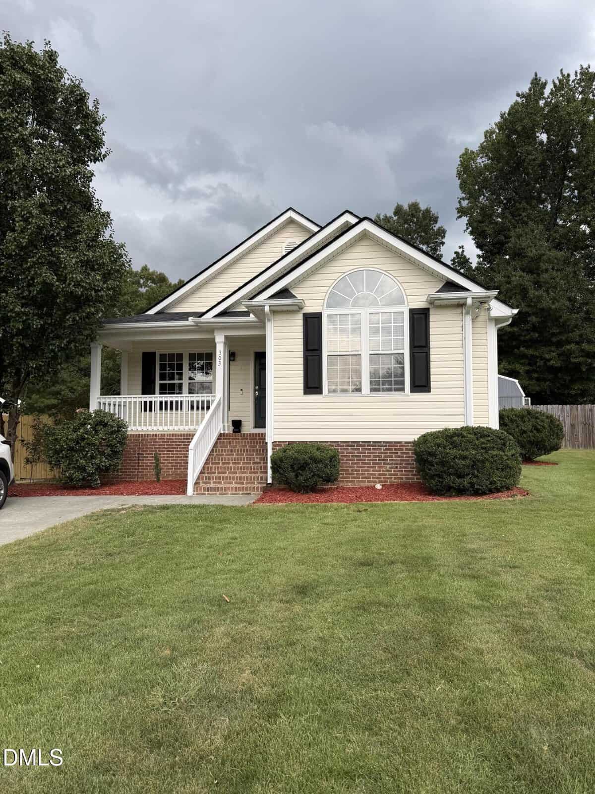 Single-story beige house with black shutters, arched window, brick foundation, front porch with white railing, and red mulch landscaping at 303 Gallop Court. A well-kept lawn and trees stem gracefully around the house under a cloudy sky.