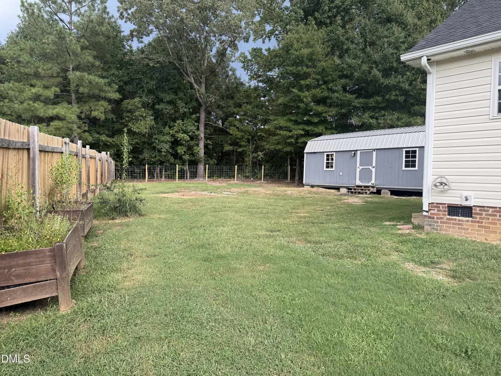 A backyard with green grass, a gray shed on the right, wooden garden beds on the left, a wooden fence, and trees in the background under a cloudy sky.