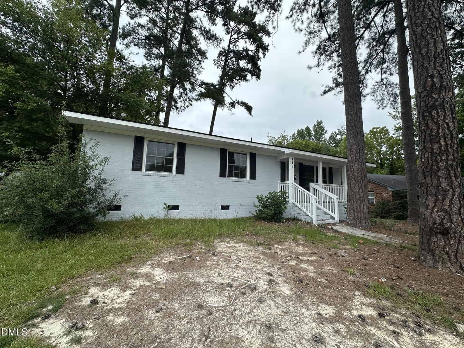 A small white brick house with dark shutters, a front porch with steps and railing, surrounded by trees and sparse grass on a cloudy day sits quietly at 113 Beam Circle, Louisburg.