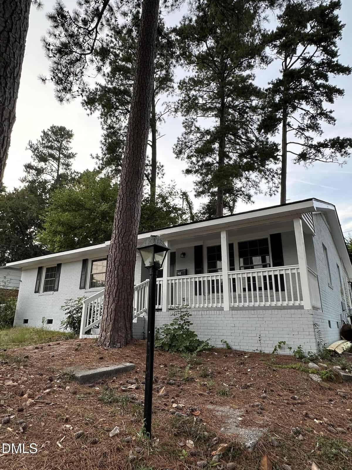 A white brick house with a covered front porch, surrounded by tall pine trees. A black lamp post stands near the front walkway, and pine needles cover the ground.