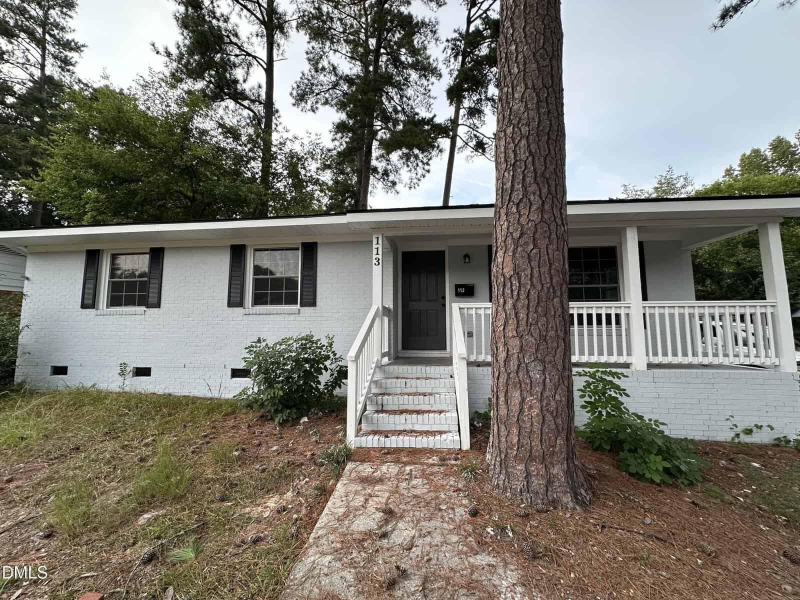A small white brick house with black shutters and front steps, surrounded by tall trees and a patch of grass and pine straw in the yard.