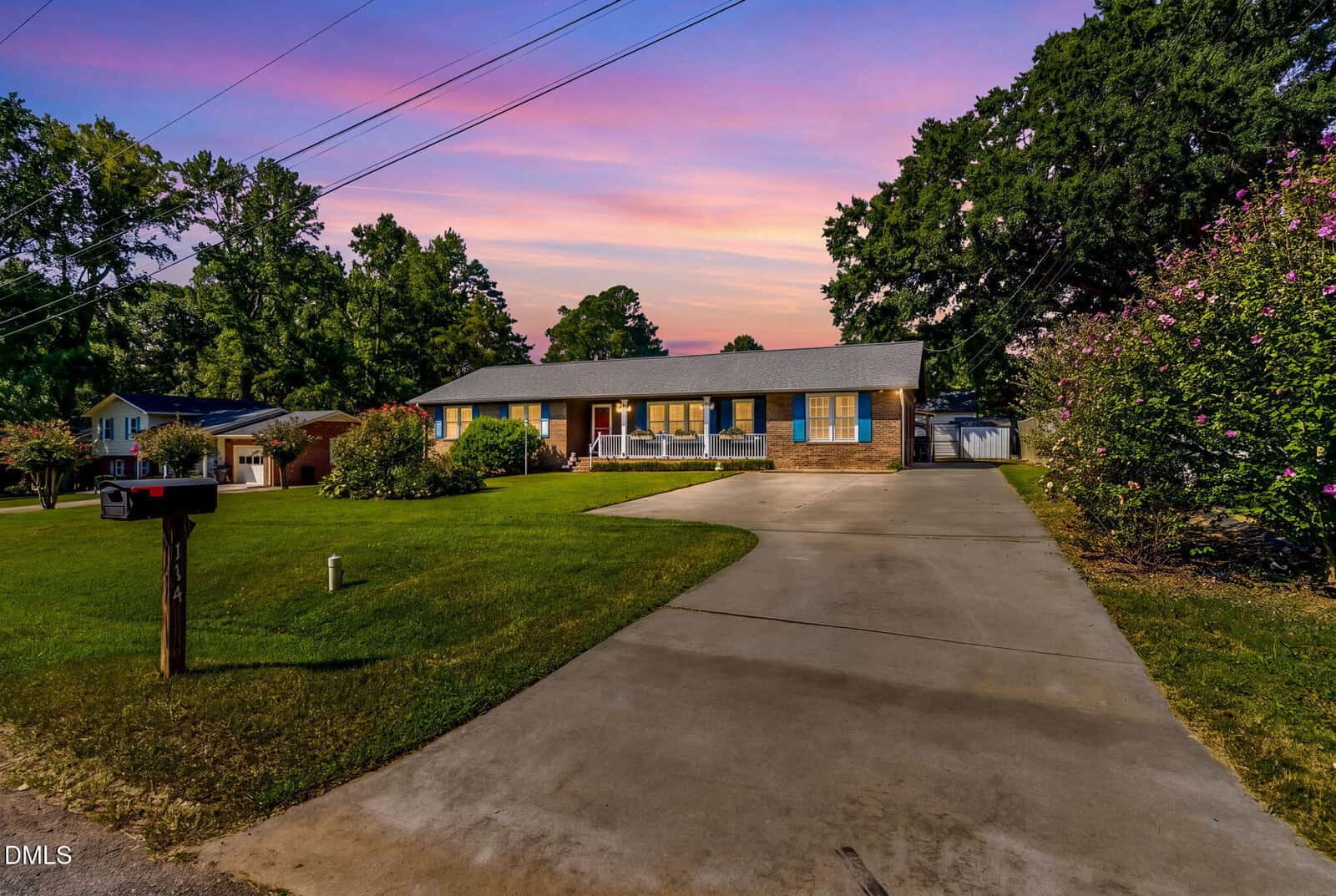 A single-story brick house with blue shutters, a wide driveway, green lawn, mailbox, trees, and a pink-purple sunset sky in the background.