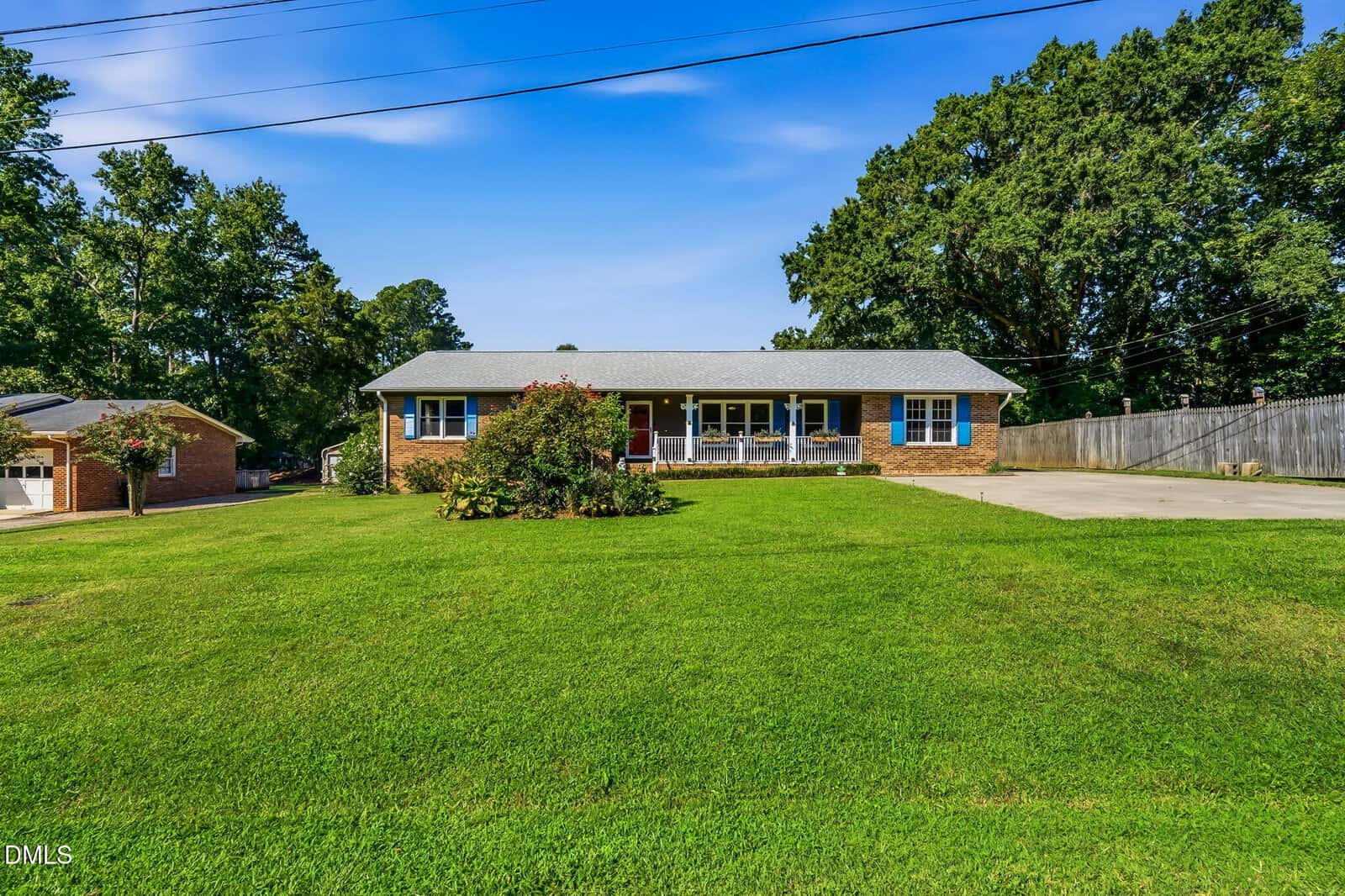 A single-story brick house with a large green lawn, a front porch with white railings, and a wide driveway, set against a backdrop of trees under a clear blue sky.