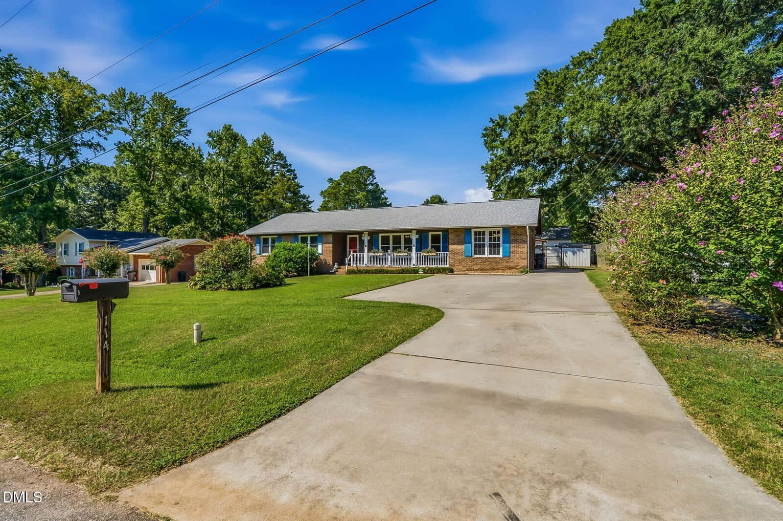 Single-story brick house at 114 Dorsey Place in Henderson with a large front yard, long driveway, welcoming front porch, and mature trees surrounding the property under a blue sky with scattered clouds.