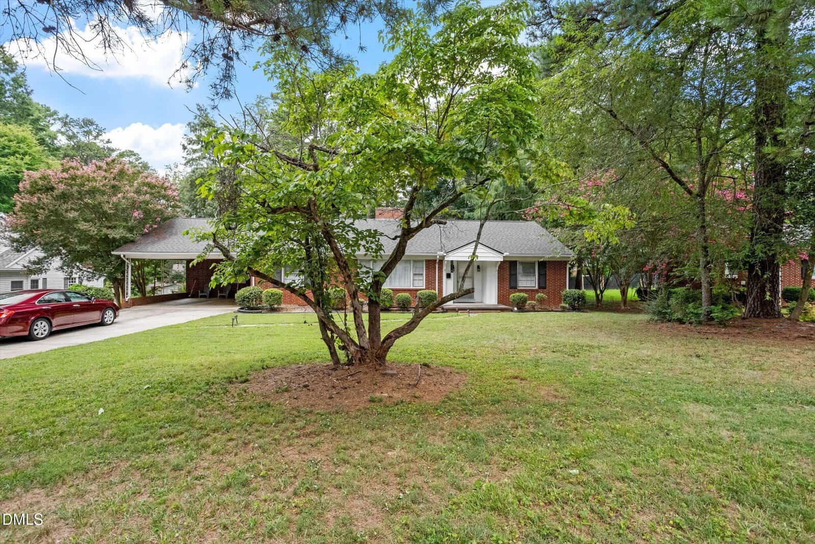 Single-story brick house with white trim, a covered carport, and a large front yard with trees and bushes located at 1516 Cypress Drive in Henderson. A red car is parked in the driveway to the left. The sky is blue with some clouds.