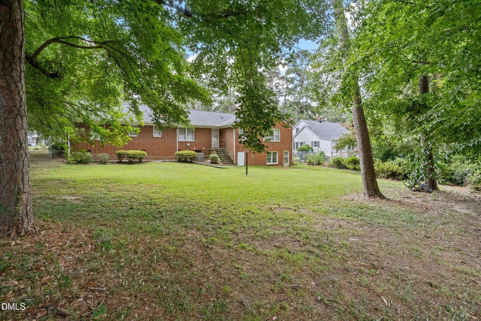 A brick house with white trim sits in a spacious, grassy backyard surrounded by tall trees. Another house is visible nearby. The area is shaded, and the lawn is well-maintained.