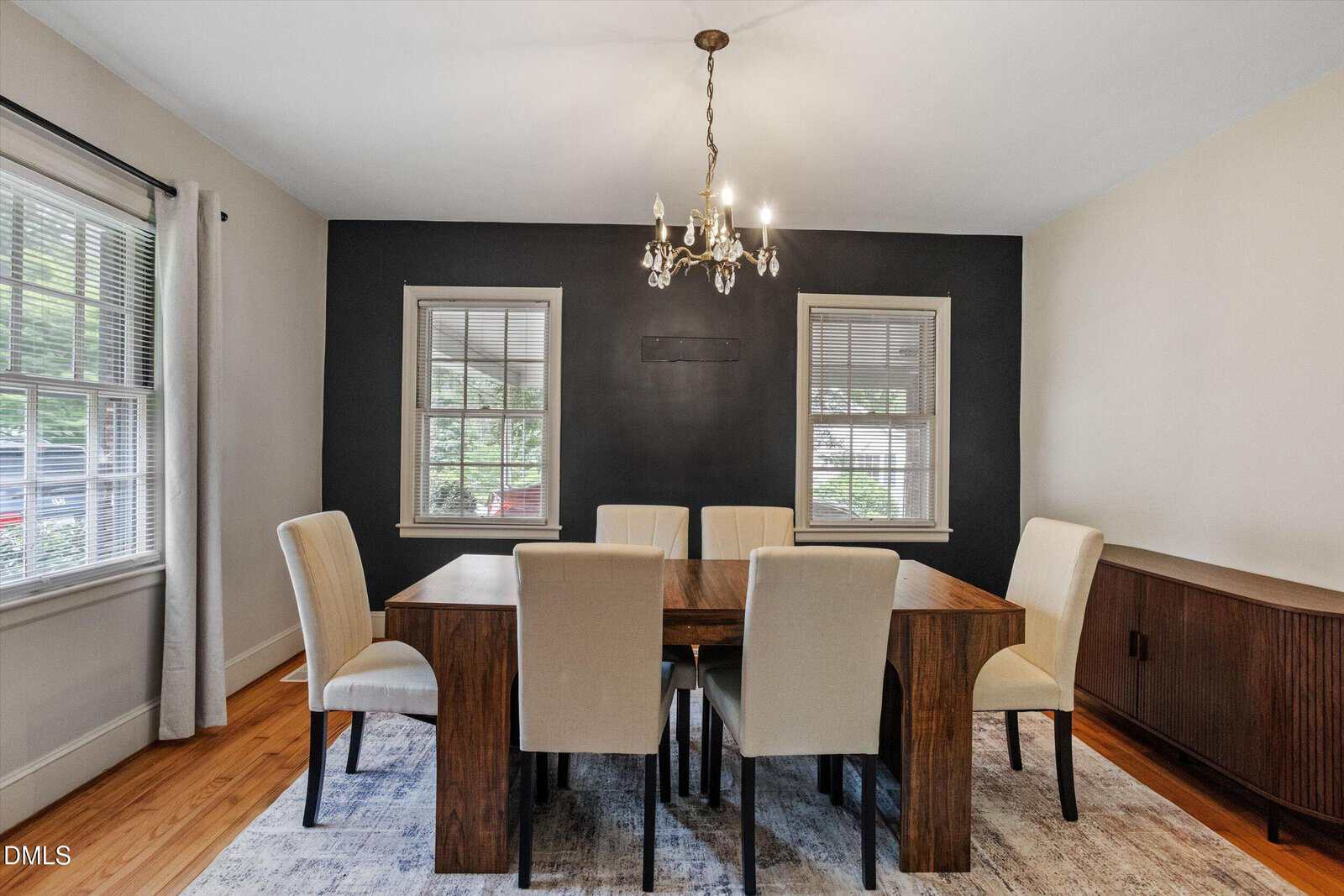 A dining room with a wooden table, six cream-colored chairs, a dark accent wall, two windows, a chandelier, and a sideboard on a light rug. Natural light enters through the windows.