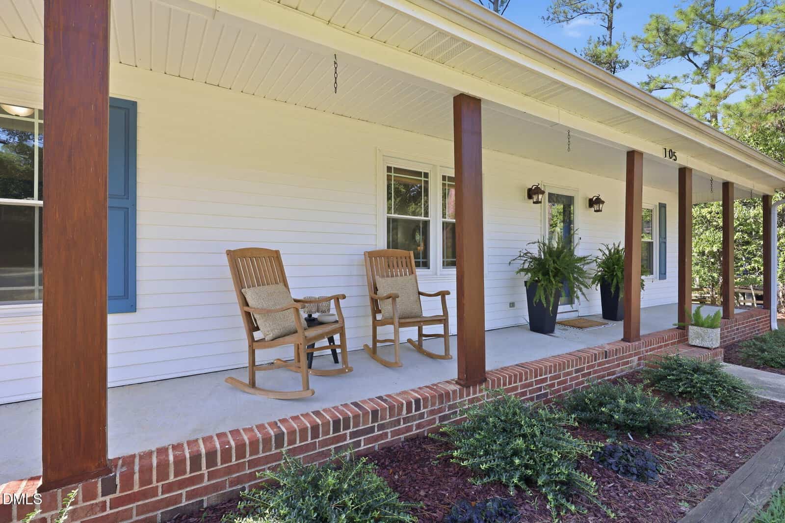 A white house with a front porch featuring two wooden rocking chairs, brown columns, potted ferns, blue shutters, and a brick-edged walkway with shrubs and mulch in the garden.
