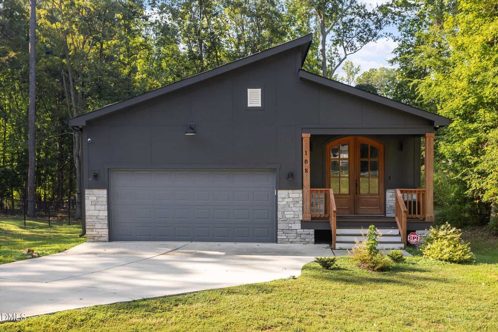 Modern dark gray house at 108 Blackfoot Drive in Louisburg with a two-car garage, stone accents, wooden porch steps, double wooden front doors, and surrounded by green trees and lawn on a sunny day.