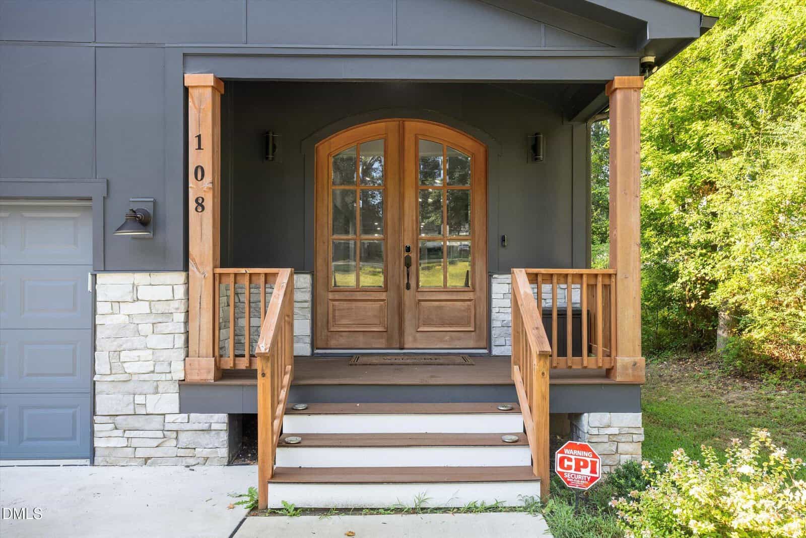 Wooden double front doors with glass panes and arched tops, flanked by wooden railings and steps. The house exterior is gray with stone accents. A CPI security sign is visible near the steps, and greenery surrounds the entrance.