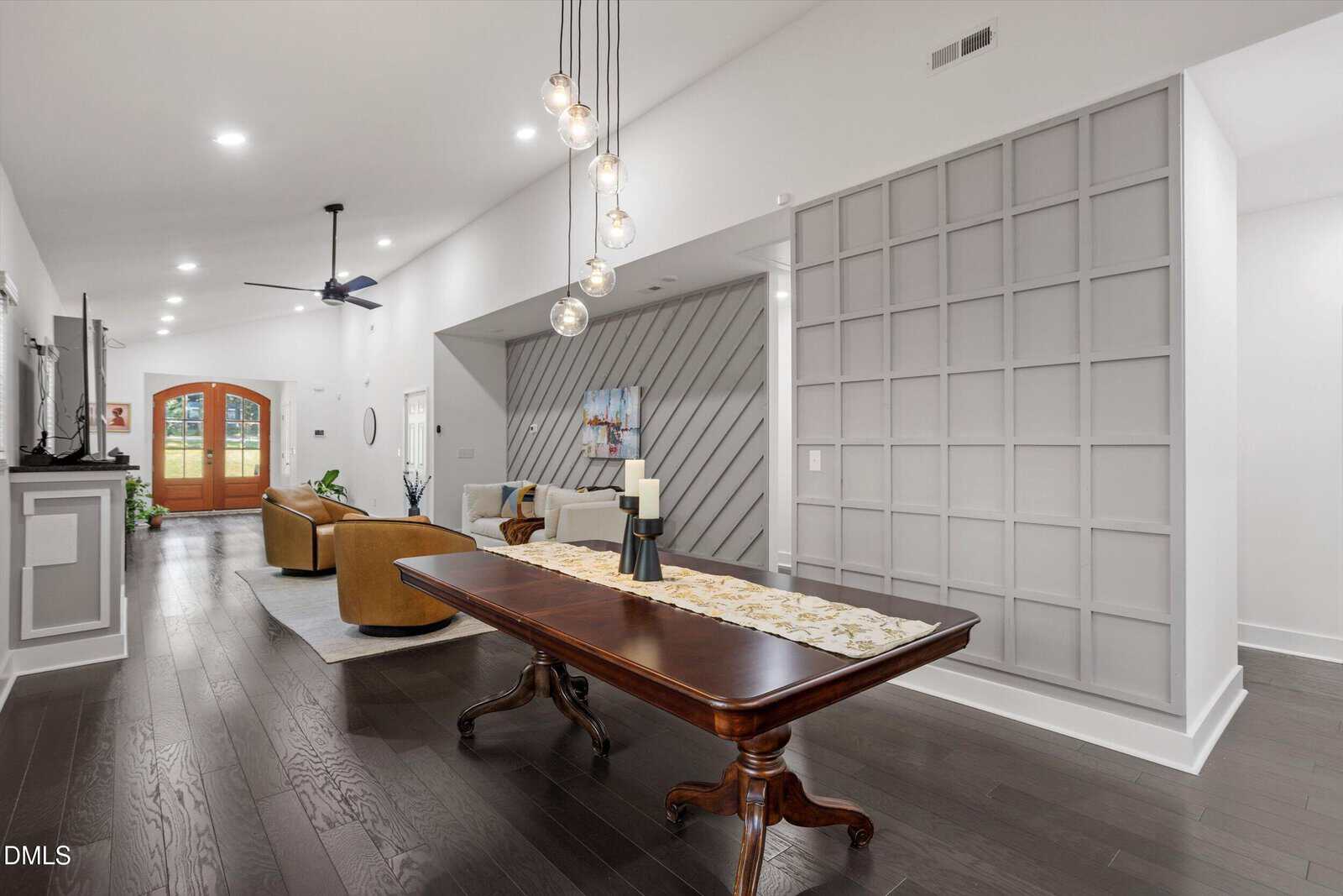 Modern open-concept living space with dark wood floors, a long wooden table, tan chairs, wall art, unique pendant lights, and large double wooden front doors with glass panels. Contemporary accent wall on the right.