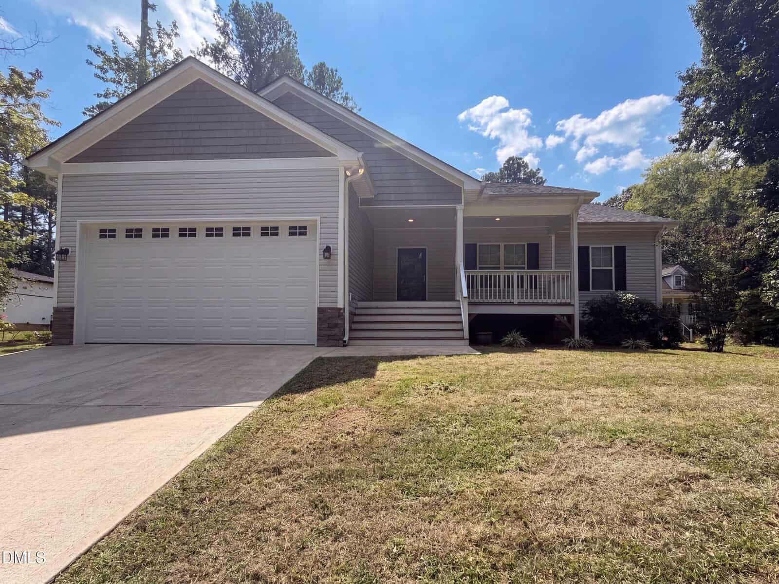 Single-story house in Louisburg with a large garage, front porch with white railings, and a sloped driveway. Located at 544 Shawnee Drive, it features light gray siding and a gable roof. Lawn and trees are visible, with a clear blue sky and a few clouds above.