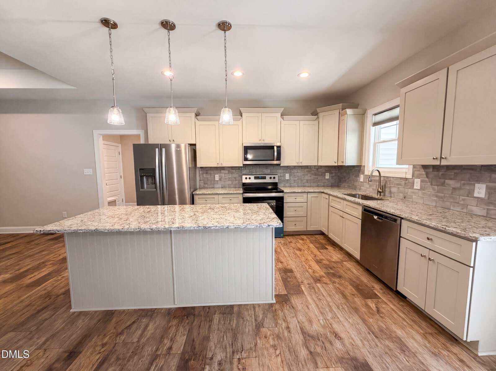 Modern kitchen with light cabinets, granite countertops, stainless steel appliances, a large island with pendant lights, tile backsplash, and wood-style flooring. Natural light comes in from a window above the sink.