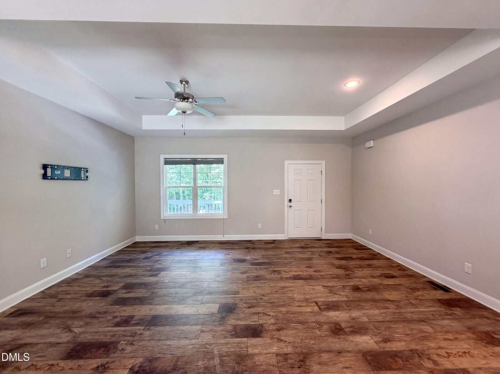 Spacious empty living room with wood floors, a ceiling fan, recessed lighting, a large window, and a white front door. Walls are light-colored, and natural light comes through the window.