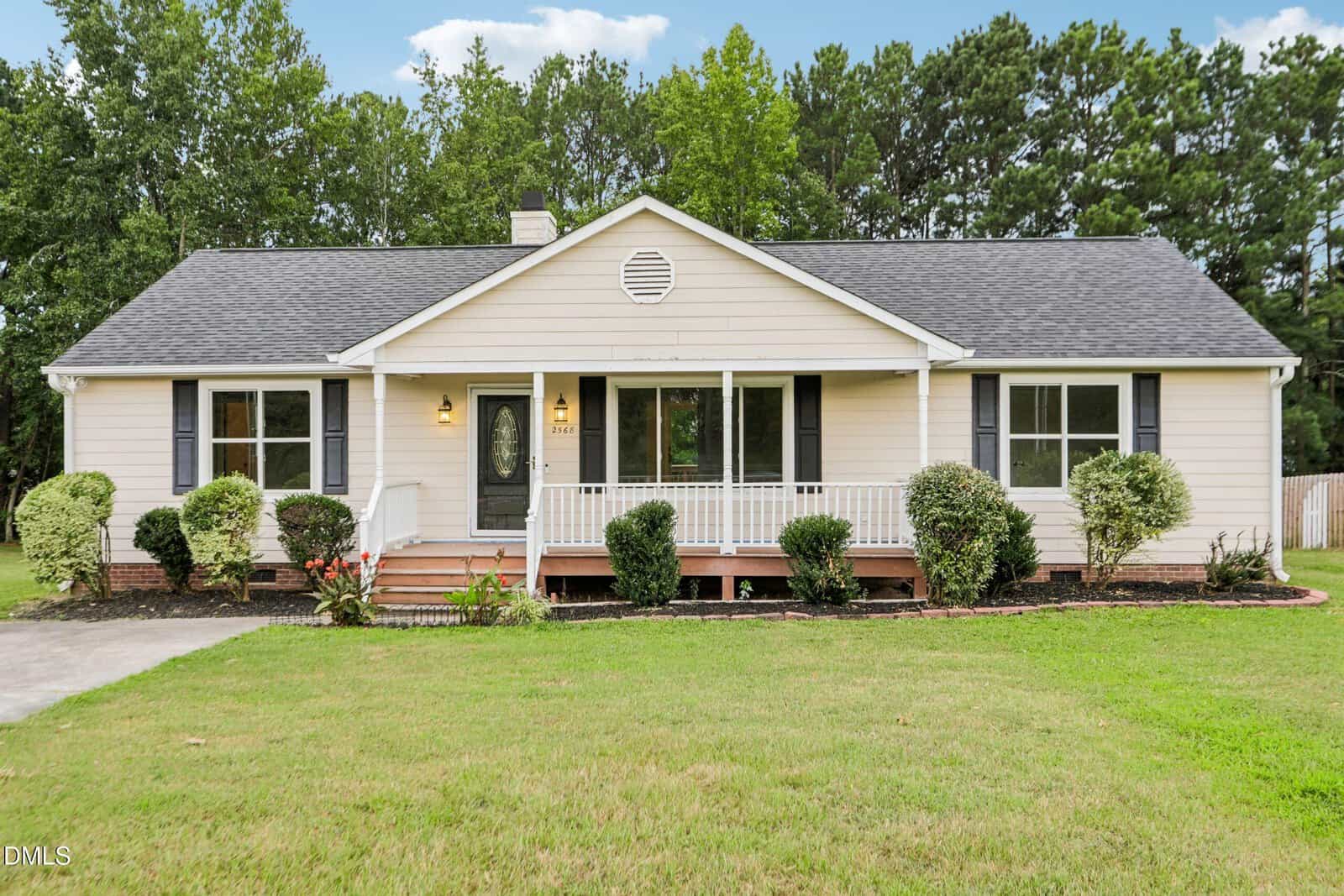 Charming single-story beige house on 2568 Wilton Avenue in Creedmoor with a black shingle roof, white trim, inviting front porch, and classic black shutters. Neatly trimmed bushes and lush green lawn grace the front, framed by mature trees in the background and a clear blue sky overhead.