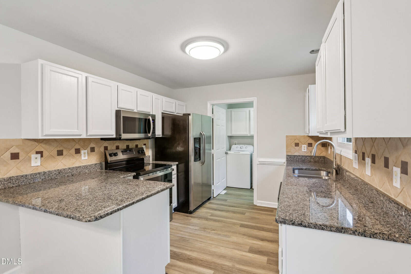 Modern kitchen with white cabinets, granite countertops, stainless steel appliances, and wooden flooring. Laundry area with washer and dryer is visible through an open door in the background.