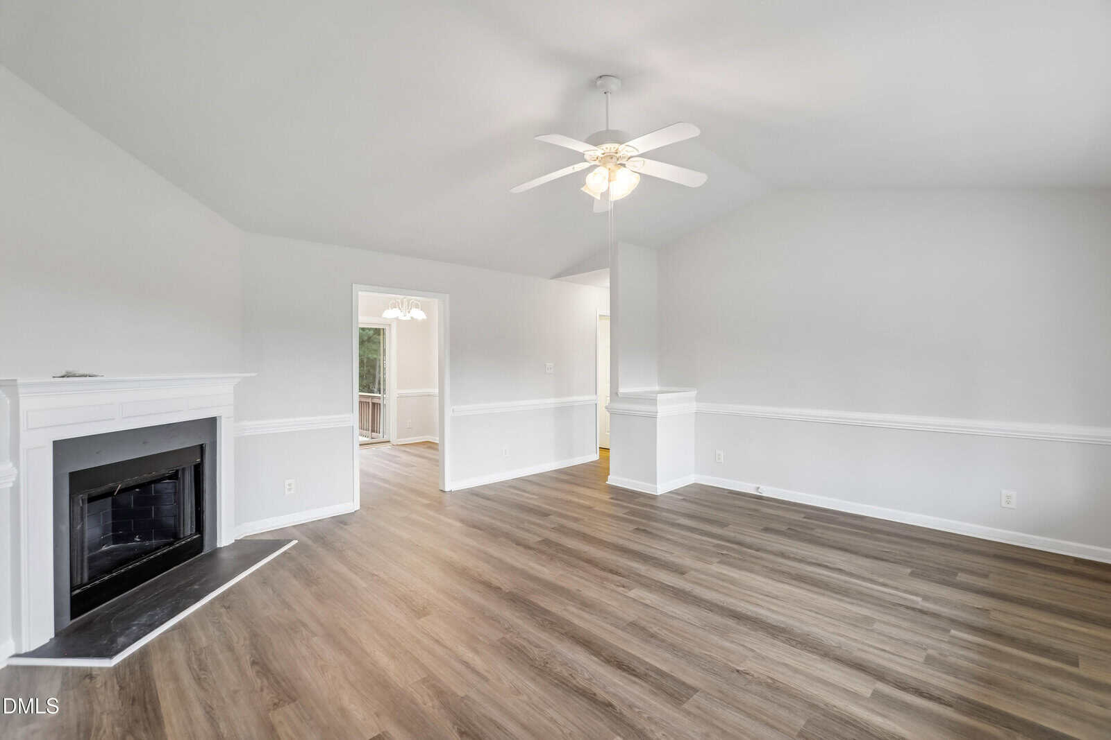 Bright, empty living room with light gray walls, wood flooring, a white ceiling fan, a corner fireplace, and an open doorway leading to a dining area with a chandelier and visible natural light.