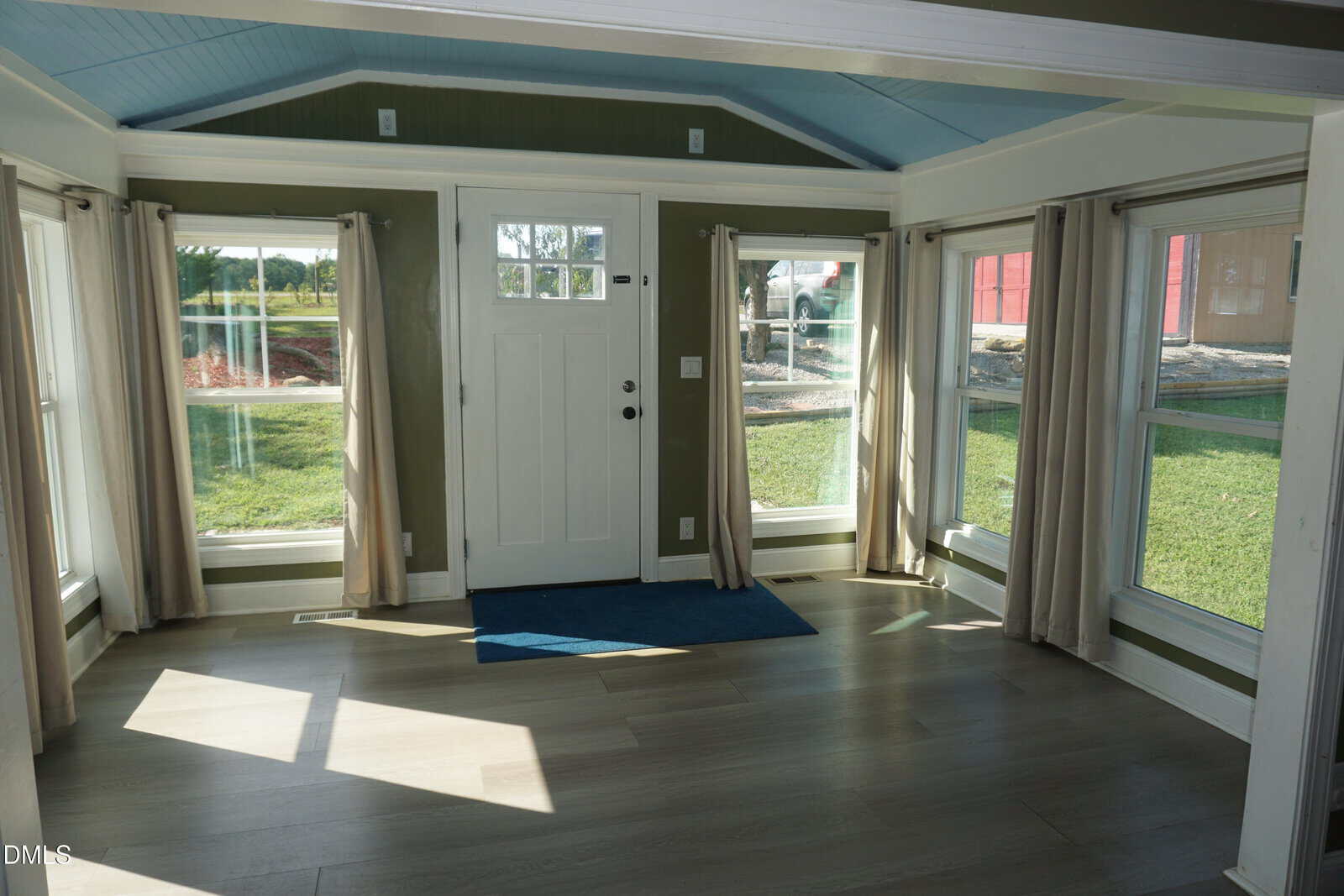 Bright room with large windows, a white door, beige curtains, light wood floors, a blue doormat, green walls, and a blue ceiling; outside, grass and a red structure are visible at 1175 Gardner Road in Castalia.