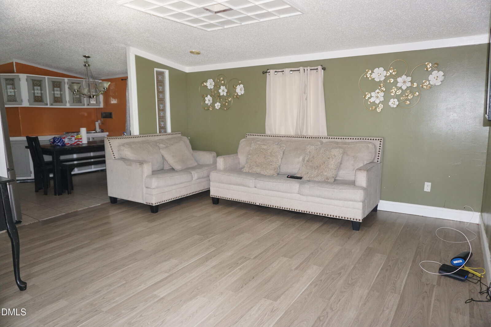 Living room with a beige loveseat and sofa, light wood flooring, green accent wall with floral decor, and a window with closed beige curtains. Dining area visible in the background. Cables are on the floor to the right.