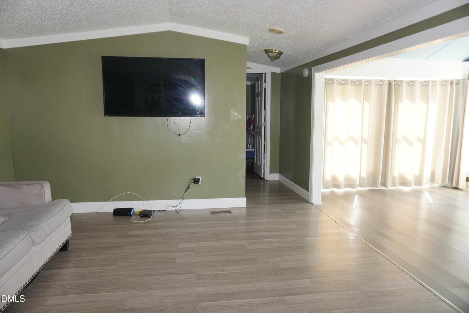 A living room with light wood flooring, a wall-mounted TV, an exposed cable, a beige couch, and an adjacent space with large windows covered by beige curtains.