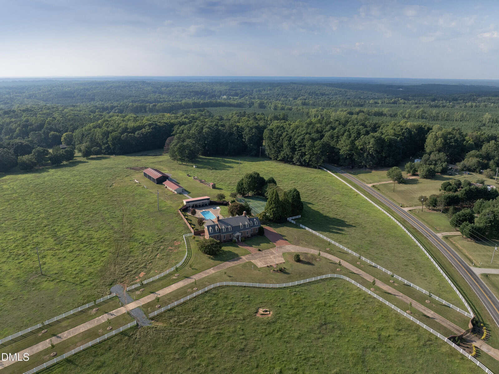 Aerial view of 32 Wes Sandling Road in Franklinton showcases a large rural property with a main house, pool, multiple outbuildings, fenced green pastures, and a winding paved driveway bordered by trees and open fields. Dense forest surrounds the property in the distance.