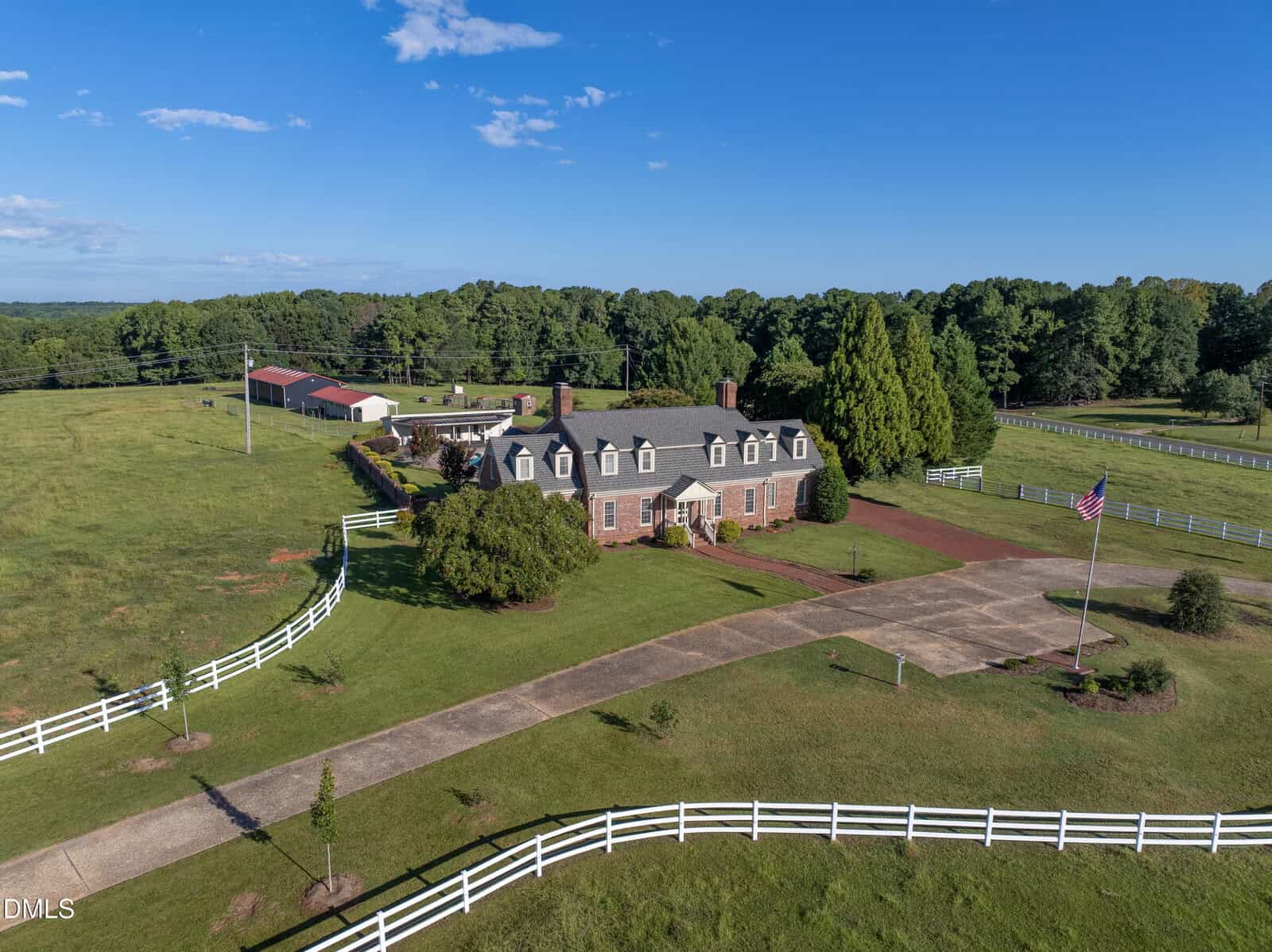 Aerial view of a large brick house with a gray roof, surrounded by a white fence, green grass, trees, a driveway, an American flag, and outbuildings under a blue sky.
