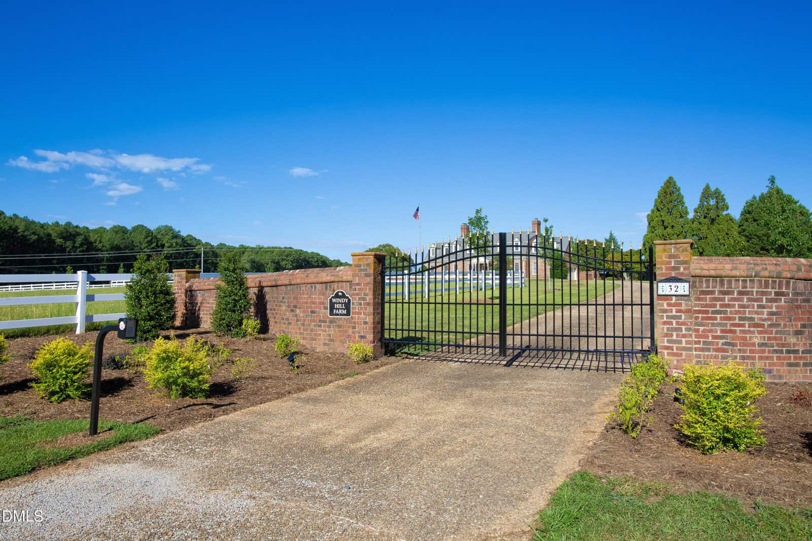 A black iron gate set in a brick wall marks the entrance to a property with manicured landscaping, a gravel driveway, and green grass under a clear blue sky.
