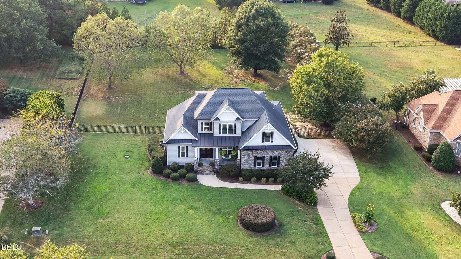 Aerial view of a two-story house with a gray roof, stone and siding exterior, circular driveway, trimmed bushes, and large green yard surrounded by trees.