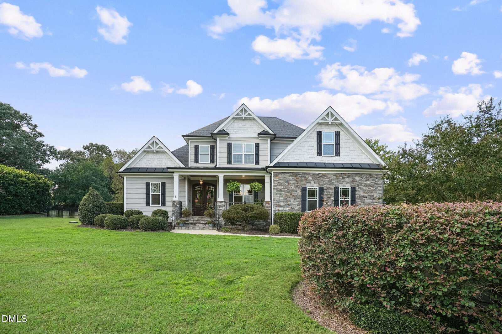 Two-story house with stone and siding exterior, gabled roof, front porch with columns, large front lawn, trimmed bushes, and trees under a blue sky with clouds.