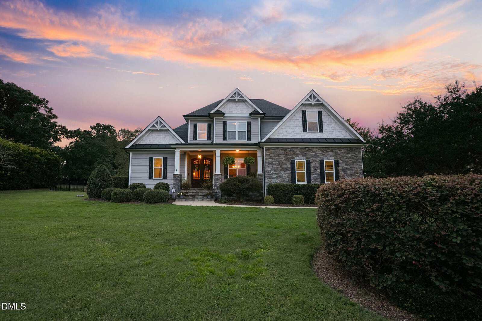A two-story suburban house with a stone and siding exterior, lit warmly from inside, sits amid a large green lawn at sunset with a colorful sky in the background. Nestled on 5909 Two Pines Trail in Wake Forest, this inviting home beautifully captures the charm of its serene neighborhood setting.
