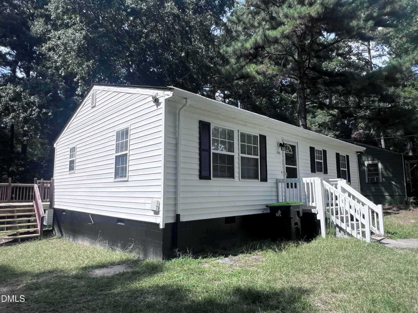 Single-story white house with black shutters, a small front porch with white railings, steps leading to the door, and surrounded by grass and tall trees.