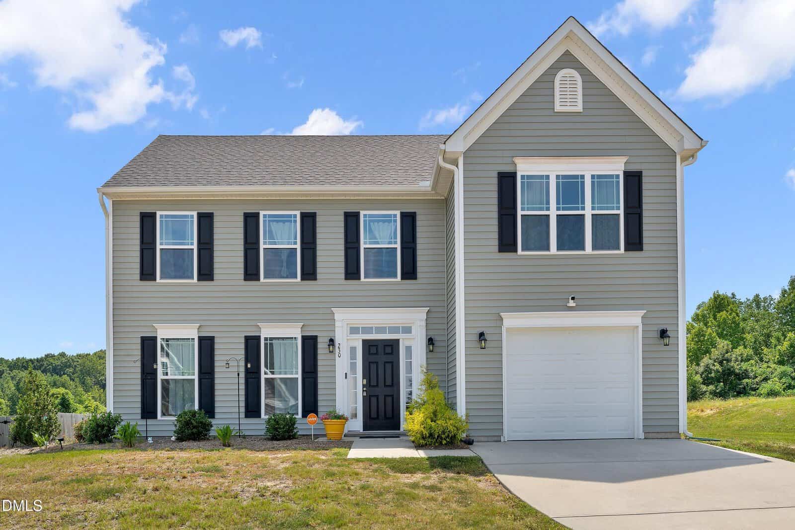 Two-story gray house with black shutters, white trim, single-car garage, and a front yard with small shrubs. Blue sky and greenery in the background.