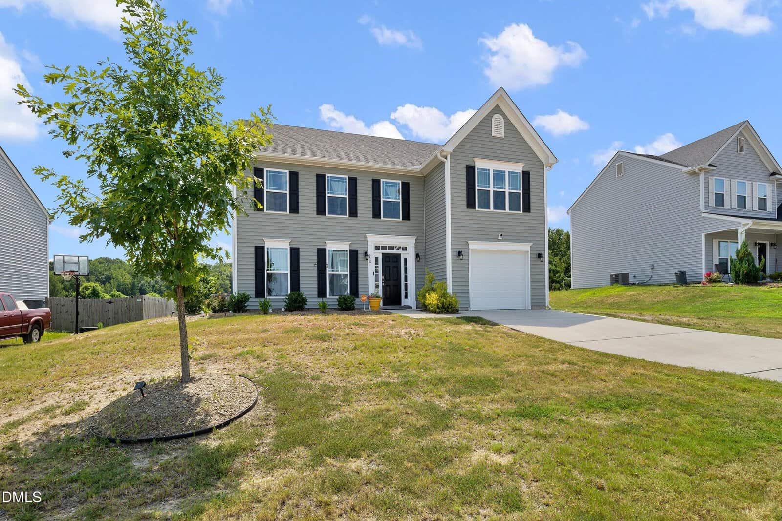 Two-story gray house with white trim and black shutters, attached single-car garage, small front porch, and a tree in the front yard. The lawn is partly brown, and neighboring houses are visible.