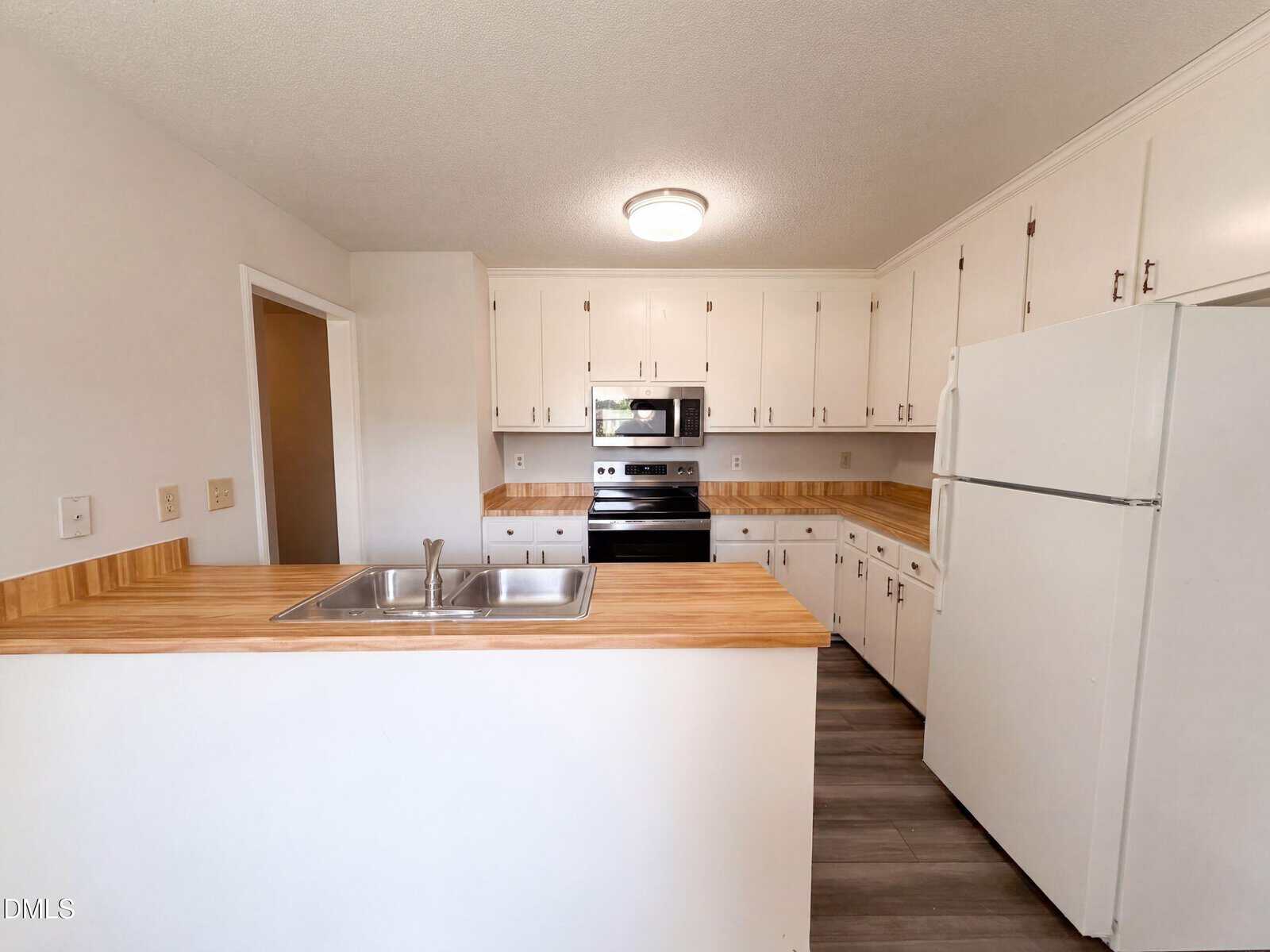 Modern kitchen with white cabinets, wood countertops, stainless steel sink, electric stove, microwave, and a white refrigerator. Light gray flooring and overhead lighting are also visible.