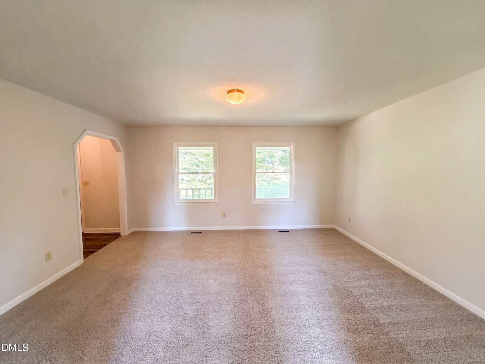 A spacious, empty living room with beige carpet, white walls, two windows, and a ceiling light. An open doorway leads to another room with wood flooring.