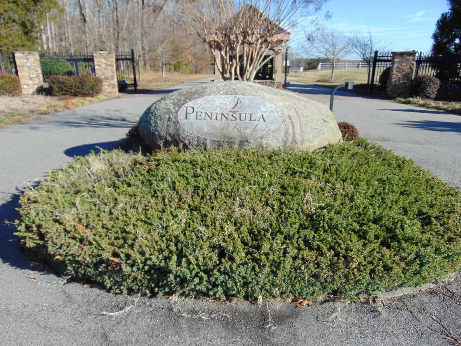 A landscaped entrance area featuring a large rock with the inscription "peninsula" resting on a bed of low-lying shrubbery, with metal gates visible in the background under a clear sky.