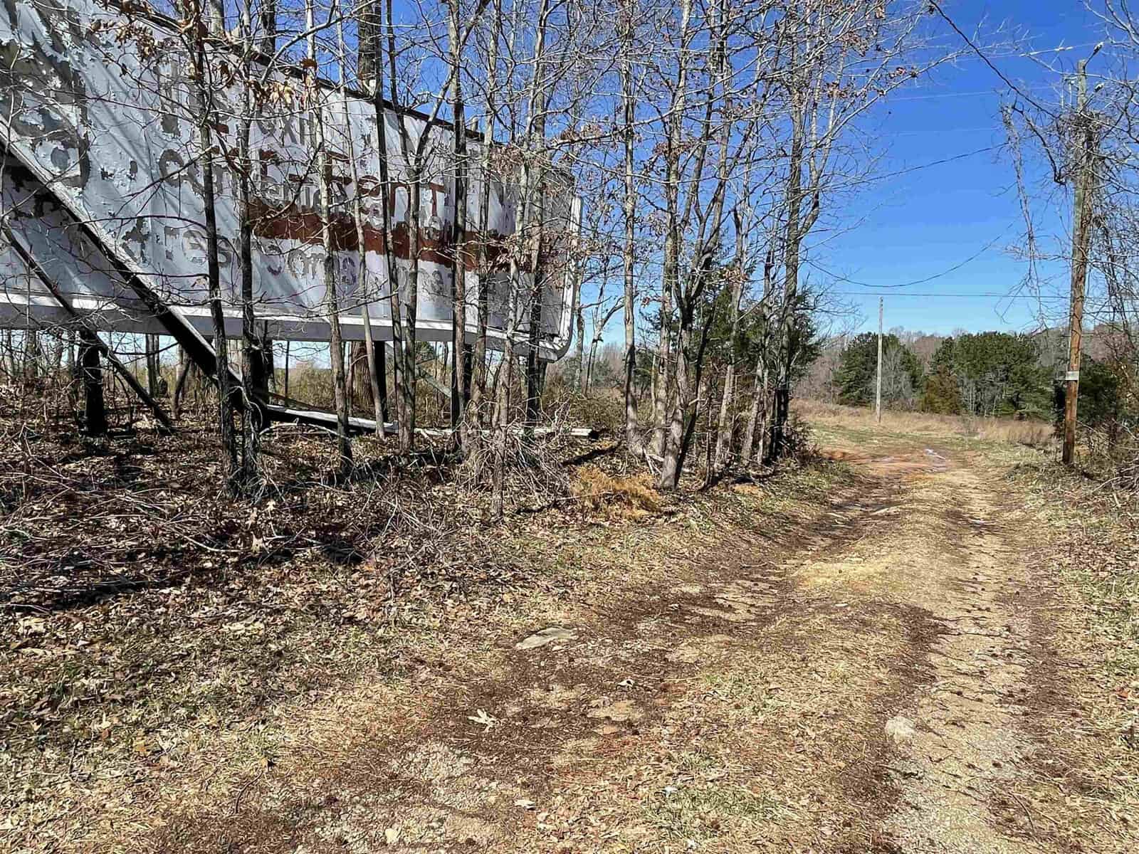 A weathered billboard stands beside a dirt path, with trees and brush surrounding the area under a clear blue sky.