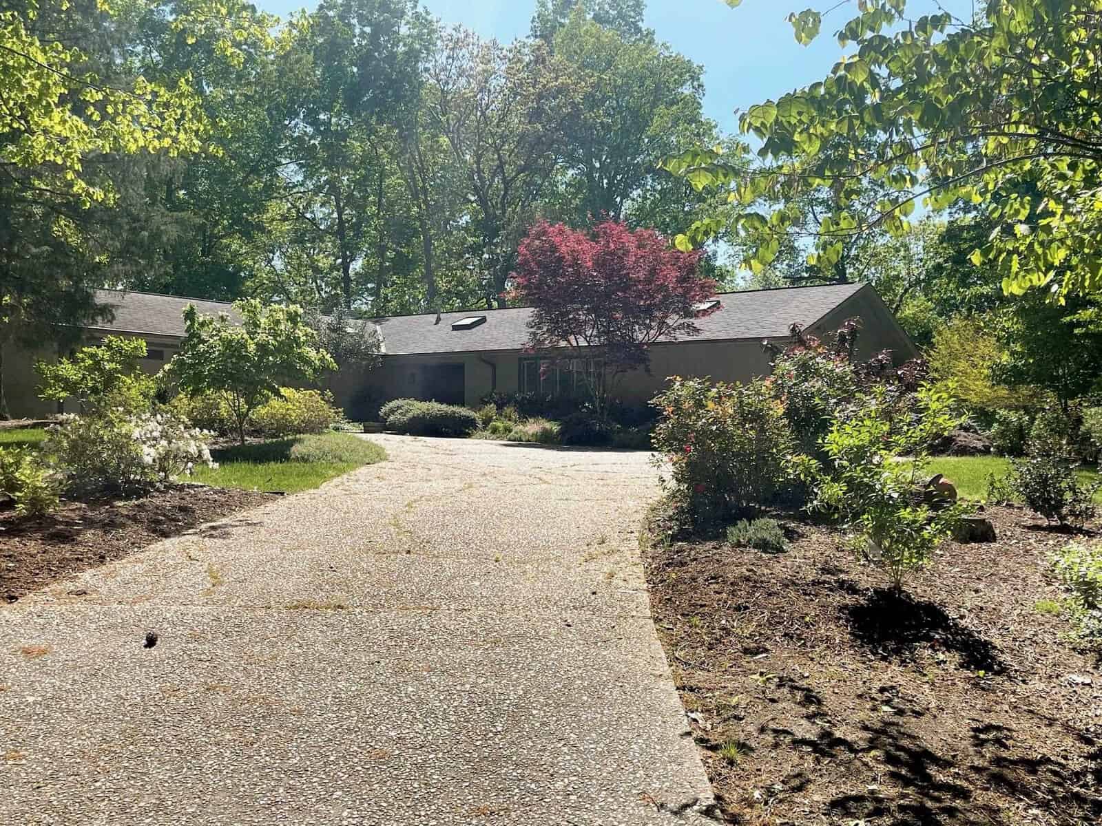 A single-story house with a red roof is partially obscured by green foliage, with a tree displaying autumnal red leaves standing out among the greenery. a driveway leads up to the home, and sunlight filters through the leaves, casting shadows on the scene.