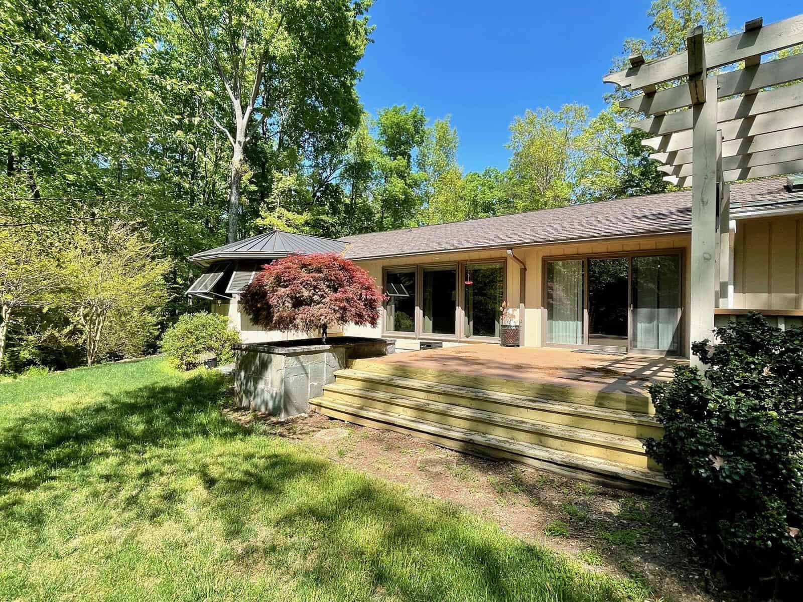 A single-story house with large windows and a wooden deck surrounded by trees on a sunny day.