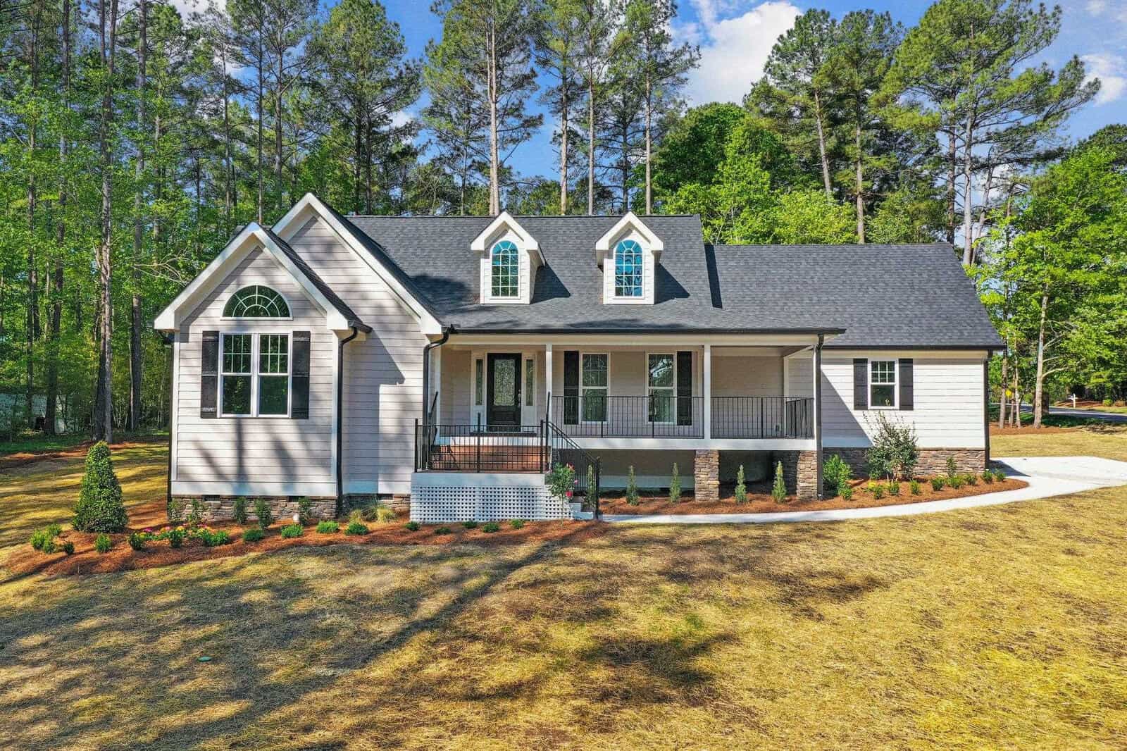 A single-story house with a front porch, gable roof, and dormer windows, set amidst a landscape with scattered trees and a freshly mowed lawn.
