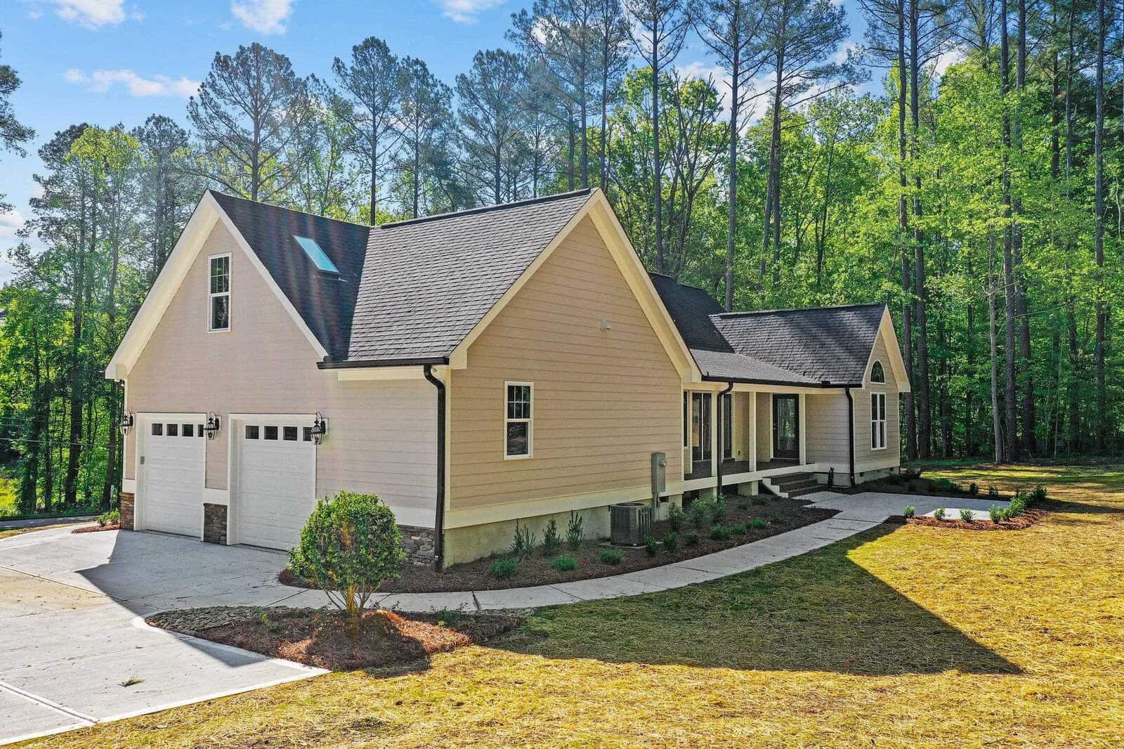 A single-story beige house with a double garage and a gabled roof, set against a backdrop of tall trees under a clear sky.