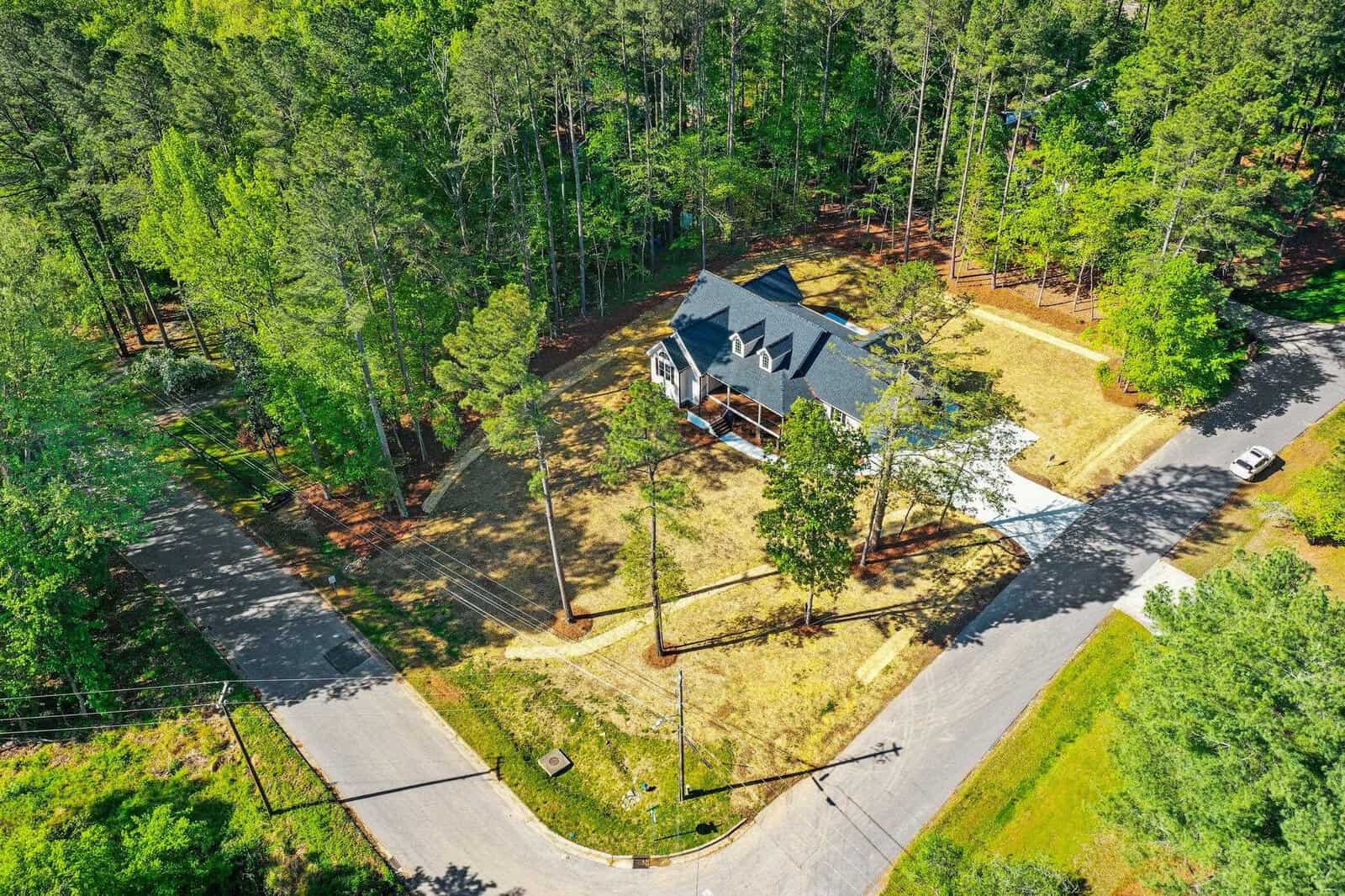 Aerial view of a single-family house surrounded by green trees in a suburban area with a clear driveway and adjacent roads.