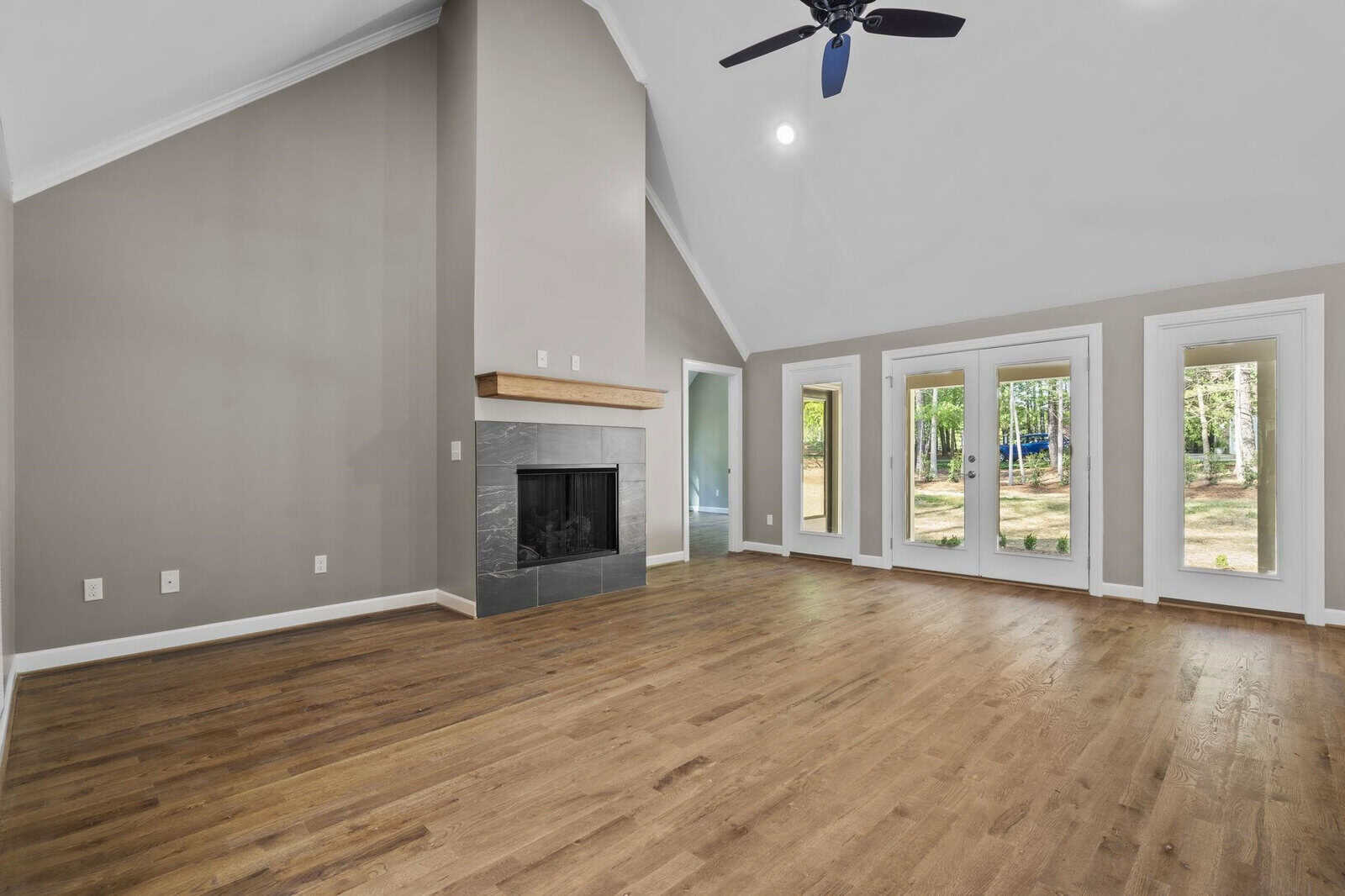 An empty living room with hardwood floors features a fireplace and multiple windows allowing natural light to enter, with views of trees outside.