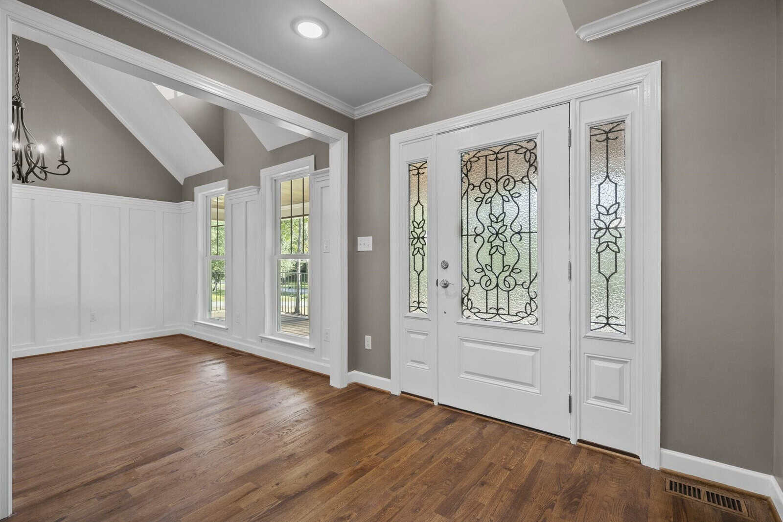 An empty room featuring polished hardwood floors, white wainscoting, and a double door with ornamental glass panes, leading to an exterior space. the room is lit by natural light from windows and has a vaulted ceiling with recessed lighting.
