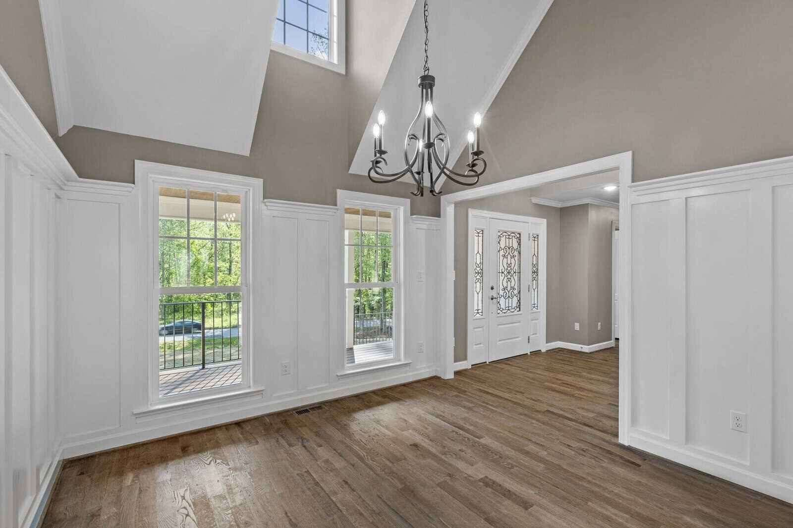 An empty room with hardwood floors, white walls with wainscoting details, multiple windows letting in natural light, and a black metal chandelier hanging from a vaulted ceiling.