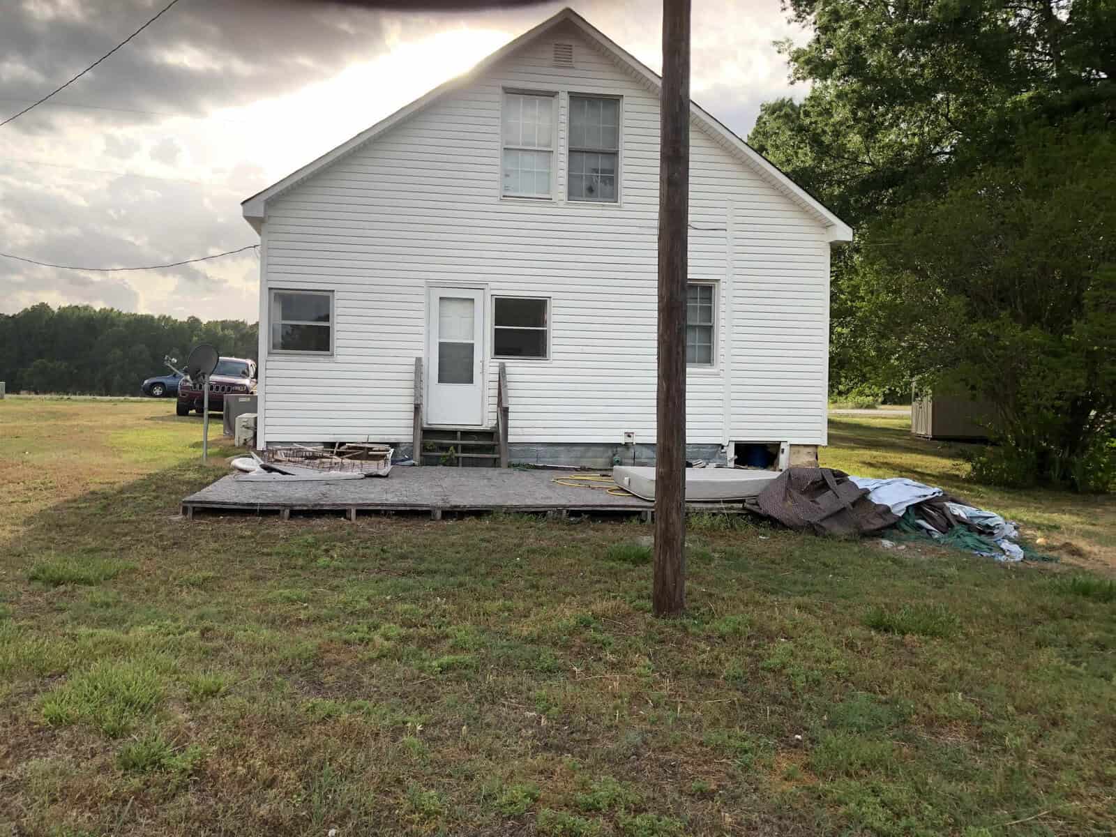 A two-story white house with a makeshift wooden porch and scattered debris on the ground, standing beside a utility pole on a grassy property.
