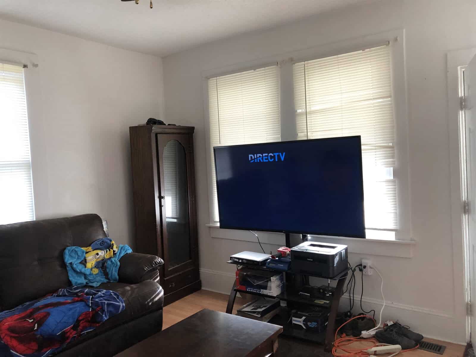 A living room with a television displaying the directv logo, a leather sofa with a blue blanket, and various items scattered on the floor.